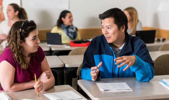 2 graduate student working together at desks with 3 other students working together in background