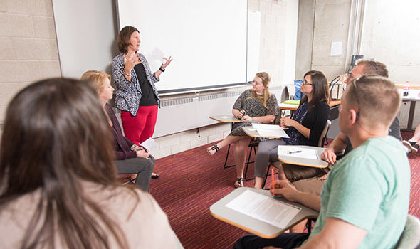 Dr. Katherine Bender standing in front of a projector screen teaching a small circle of students in desks around her