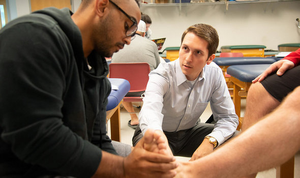 photo of Athletic Training class with 1 student sitting on a medical table, another student holding and looking at the first student's foot and a professor showing the students something on the foot