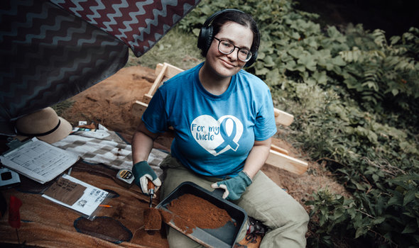 a BSU student at an archaeology dig sitting on the ground holding a dustpan and digging tool both filled with soil surrounded by a specimen bag, measuring tape, hat and clipboard