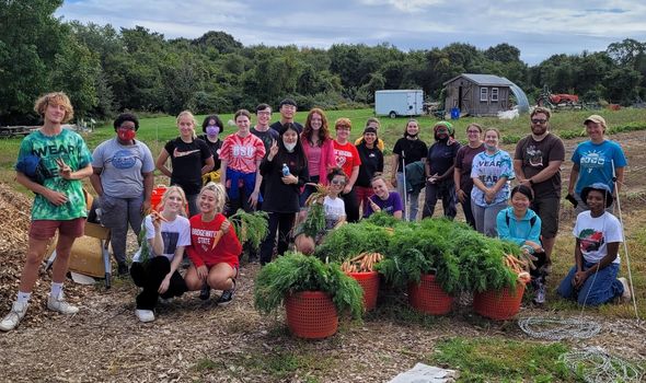 Photo of BSU volunteers at Sharing the Harvest community farm with looking at the camera and smiling. There are two rows of individuals with the back row standing and the front row crouching. In front of them are baskets of food they harvested during the service project.