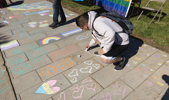 A student draws on the sidewalk with chalk; the sidewalk is decorated with hearts and rainbows for National Coming Out Day