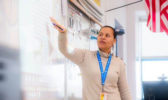 An elementary school teacher gesturing toward the whiteboard in the classroom