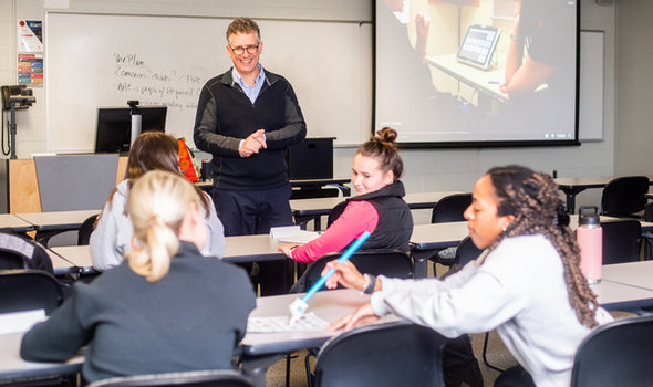 Dr. James Feeney smiling and teaching at front of class in front of whiteboard and projector screen while students in foreground practice using tools