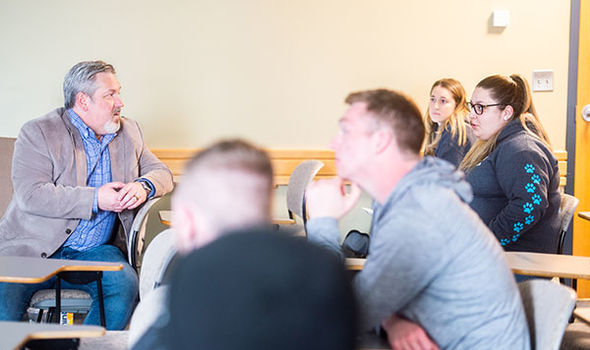 Professor Michael DeValve sitting in a classroom desk talking with 4 students in his class