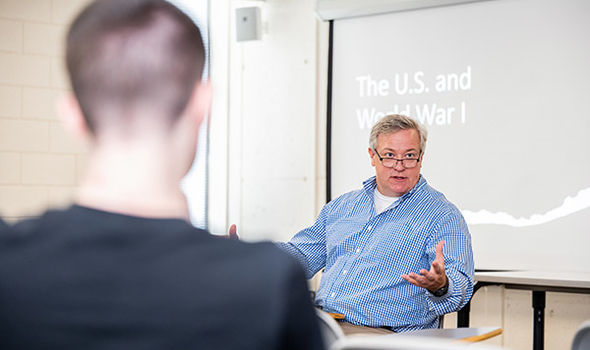 Dr. Thomas Nester sitting at the front of a classroom teaching a small group of students in front of a projector screen that says The U.S. and World War I