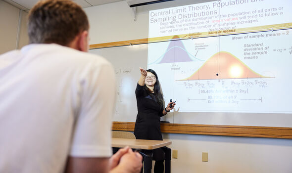 Dr. Xiangrong Liu with long dark hair, black rim glasses wearing a black collared dress smiling and gesturing in front of a white board with a projection about Central Limit Theory