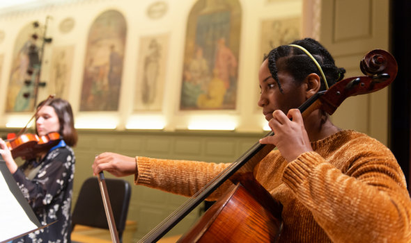 two female members of a string ensemble playing on stage; one in foreground on cello and one in background on violin
