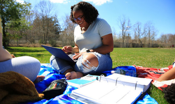 a student smiling and using her laptop computer while sitting on a blanket on the ground on campus