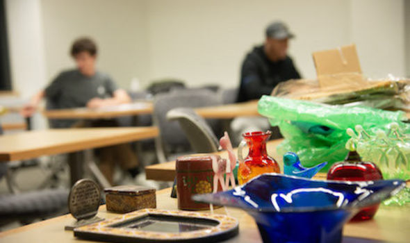 Glass art pieces sit in the foreground; in the background, students are in class