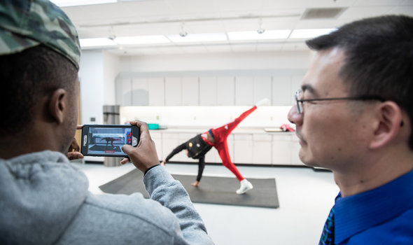 A student and professor looking through a camera to another student doing a cartwheel