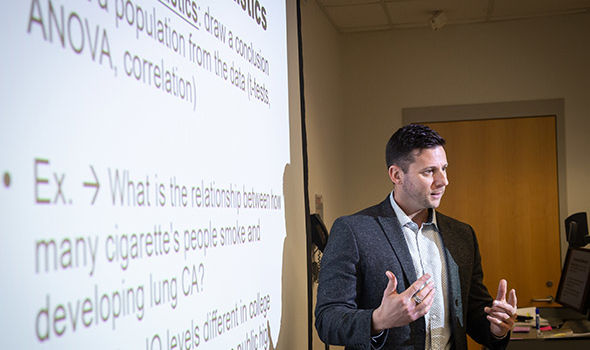 Dr. James Leone teaching a research class standing in front of a projector screen that shows an example of using statistics to correlate relationship between how many cigarettes people smoke and developing lung cancer