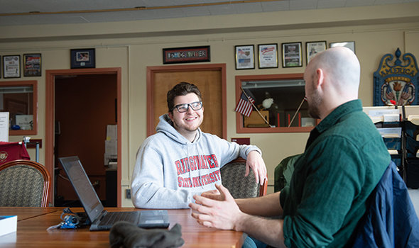 a BSU student smiling and sitting at a table while talking with a faculty member who has his laptop open between them