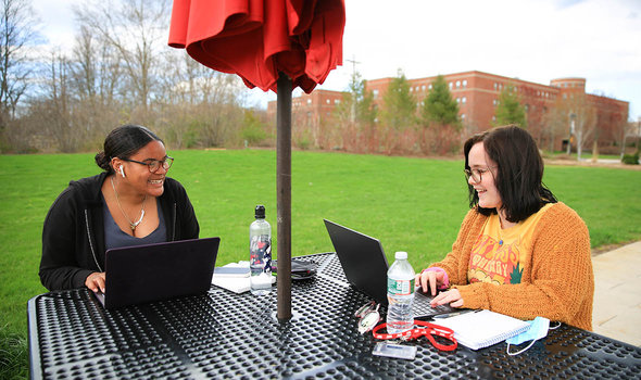 Two students sitting at a table outside, working on laptops