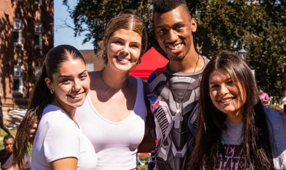 4 BSU students smiling together on the BSU quad with other students sitting or walking behind them