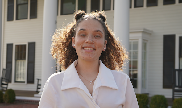 a female student with light brown, curly hair styled in space buns smiles at the camera. She is wearing earrings and a white collared blouse.