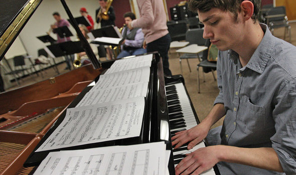 male student playing piano with 5 other students playing brass instruments in background