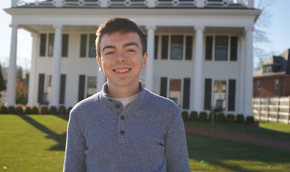 a man stands in front of a house with a medium grey 1/4 button sweater.