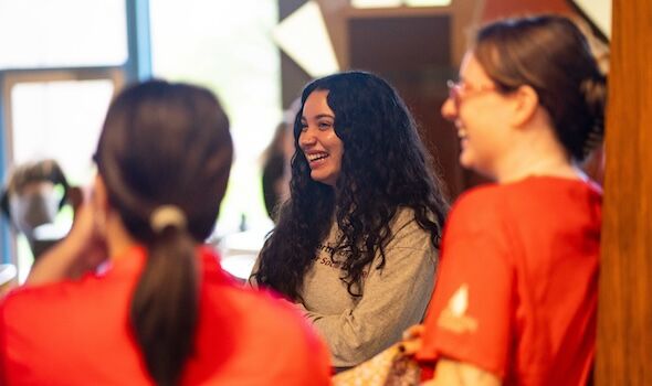 three female students stand in bears den talking and laughing. They are wearing red and grey BSU shirts respectively