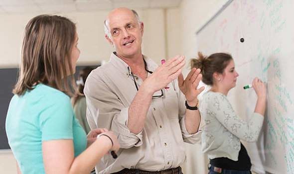 Professor Ed Carter talks to a female student in front of a classroom white board while another student writes on the board