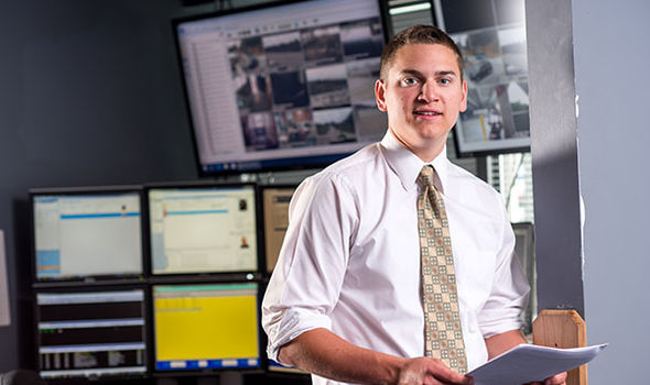 Criminal Justice student at internship with a bank of screens behind him while he stands holding a packet of papers