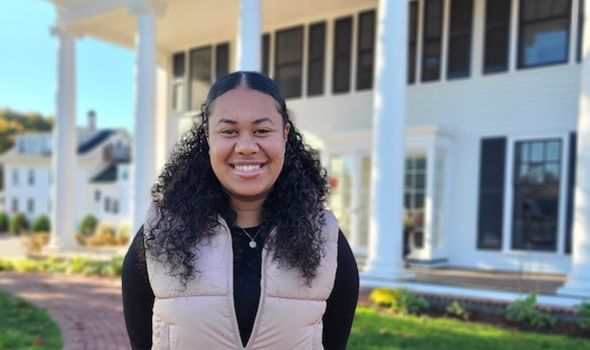 a female student smiles at the camera in front of 161 summer st. She has shoulder-length, curly black hair tied back and is wearing a black shirt with a tan vest over it and a small necklace