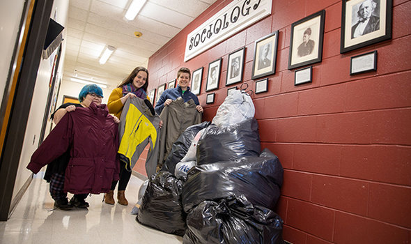 BSU Sociology professor and 2 students in hall outside Sociology office hold up coats collected for donation with bags of donations beside them on the floor