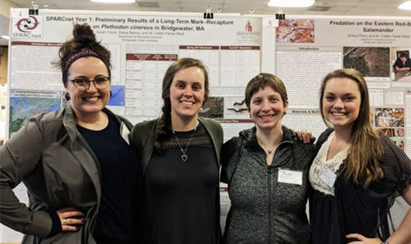Four biology undergraduate research students standing in front of their presentation