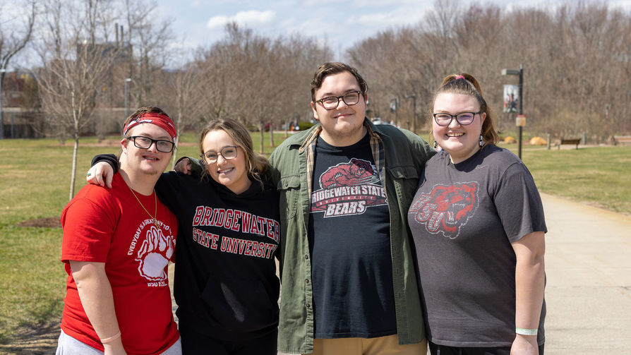4 BSU students wearing different BSU shirts stand side by side in University Park smiling at the camera