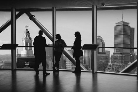 The silhouette of three people stand in front of large window with buildings in the background