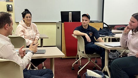 Three students and a professor talk while sitting at tables in a classroom.