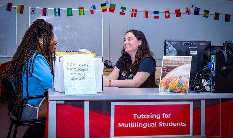 2 people sitting across from each other talking at a desk with a sign on it that says Tutoring for Multilingual Students