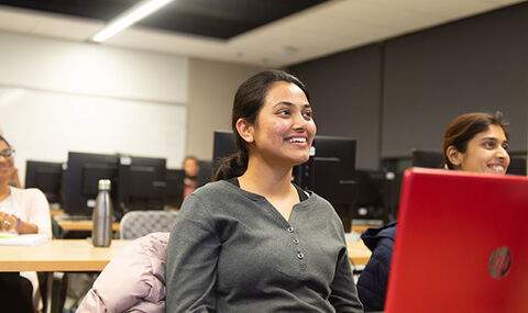 students sitting at tables in classroom with their laptops open looking at front of class