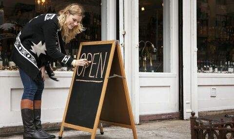 a young woman writes OPEN on a sidewalk chalkboard in front of a business