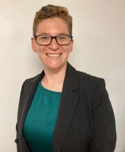 Dr. Breana Bietsch smiling with short wavy light brown hair wearing black rim glasses and a green blouse under a black blazer