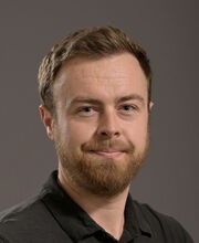Dr. Myles Bittner smiling with short reddish brown hair, mustache and beard. He is wearing a black polo shirt