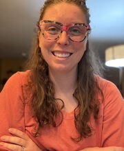 Tori Cameron smiling with long wavy light brown hair and wearing print rim glasses and an orange v-neck top