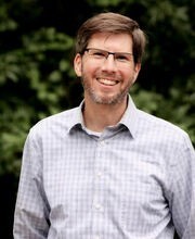 Ben Carson with short brown hair graying at the temples, brown and gray mustache and beard wearing glasses and a light blue button down shirt