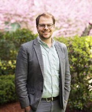 Dr. Bradford Dumont standing in a garden smiling with short dirty blonde hair, mustache and short beard. He's wearing black rimmed glasses, a gray blazer over a light green and white checked button down shirt and gray pants