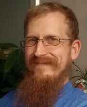 headshot photo of Stephen Flood with short reddish brown hair, mustache and beard wearing metal rimmed glasses and a blue jersey