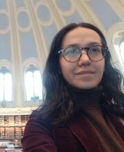 a selfie photo of Dr. Carolina Freitas inside an architectural dome decorated in a light blue, cream and gold design. Dr. Freitas has long dark brown wavy hair and is wearing brown rim glasses and a cranberry colored corduroy blazer over a brown turtleneck top.