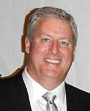 Tim Geers smiling with short gray hair and wearing a black suit jacket over a white button down shirt with a silver tie