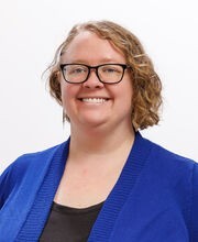 Ashley Hansen-Brown smiling with short wavy light brown hair and wearing black rim glasses and a royal blue sweater over a black blouse