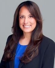Christina Hodel smiling with long wavy dark brown hair and wearing a navy blazer over a light blue blouse