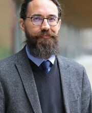 Dr. Kevin Kalish smiling slightly with dark brown hair, beard and mustache with a little gray. He is wearing a gray blazer over a black sweater vest, a light blue button down shirt and navy blue tie
