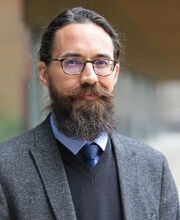 Dr. Kevin Kalish smiling slightly with dark brown hair, beard and mustache with a little gray. He is wearing a gray blazer over a black sweater vest, a light blue button down shirt and navy blue tie