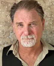 Thomas Kee with short graying dark brown hair and gray mustache and goatee. He's wearing a black vest over a beige button up shirt with the top 2 buttons undone