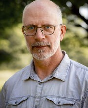 John Kucich balding with short gray hair, mustache and beard and wearing a light blue button up shirt with 2 front pockets
