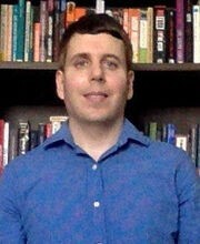 Brian Lepine with short brown hair and blue eyes wearing a blue button down shirt and standing in front of a bookcase