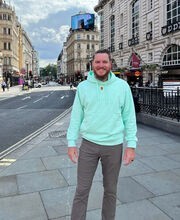 Dr. Andrew Miller standing on a city sidewalk smiling with reddish brown short hair, mustache and beard wearing a light blue green hoodie sweatshirt and gray pants